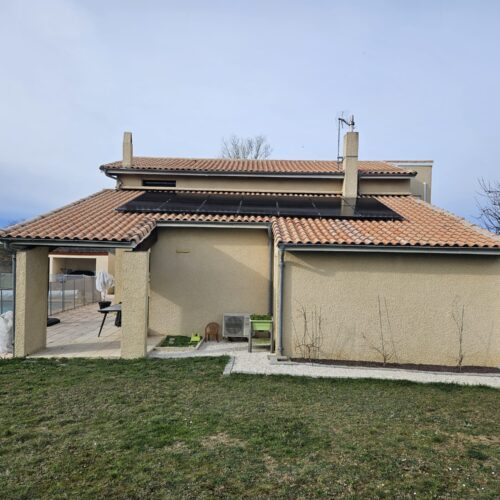 Façade d’une maison avec toit en tuiles, cheminée centrale et panneaux solaires sur la toiture, vue depuis le jardin, ciel bleu clair.