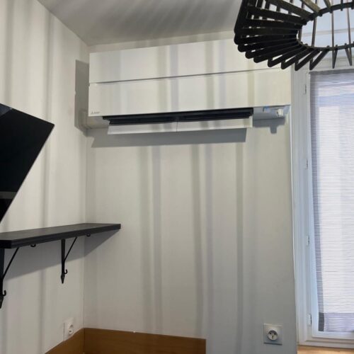 Climatisation murale blanche dans une pièce, avec étagère noire, petit bureau en bois et fenêtre à volets à droite.