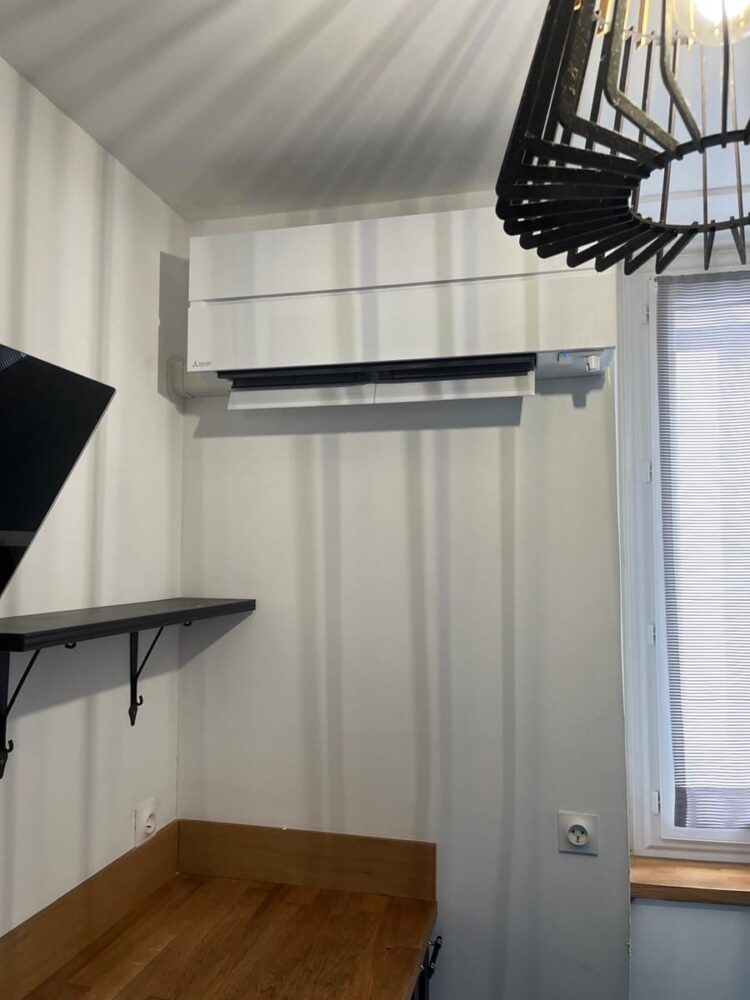 Climatisation murale blanche dans une pièce, avec étagère noire, petit bureau en bois et fenêtre à volets à droite.
