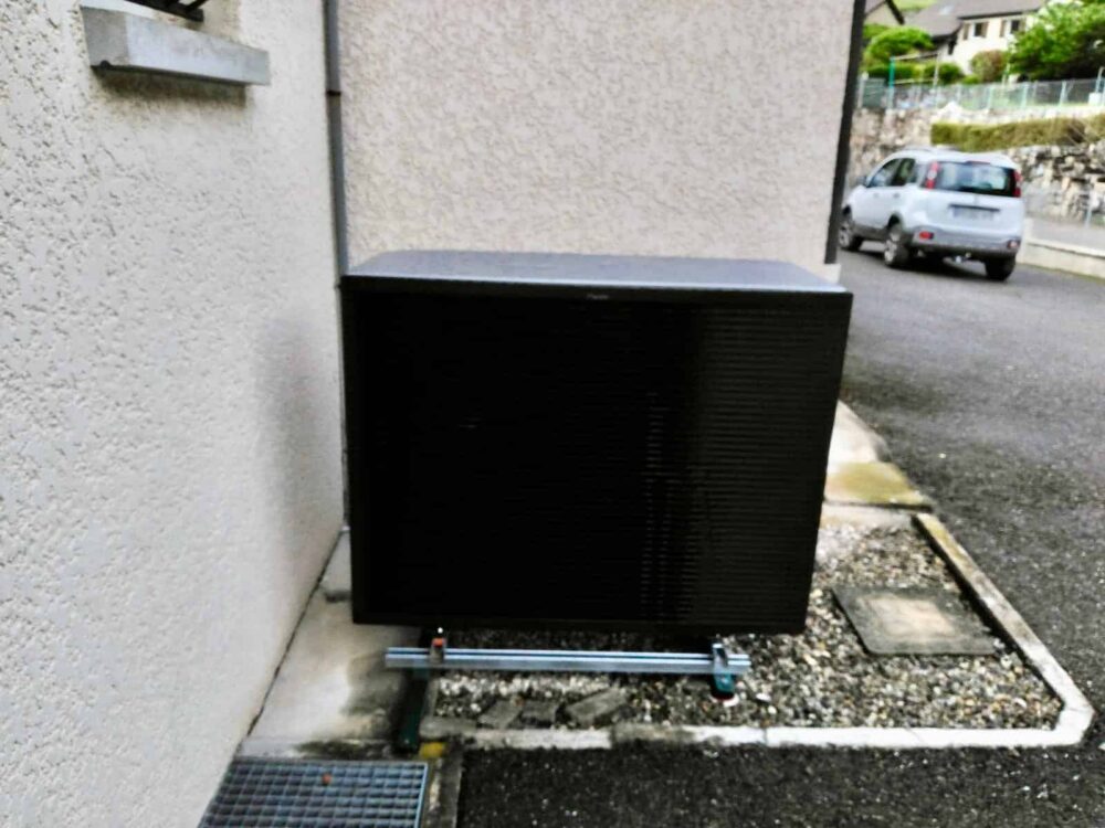Boîte noire rectangulaire sur un petit socle, installée contre un mur, dans une cour, avec une voiture garée à l’arrière-plan.