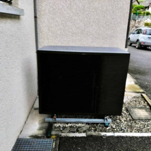 Boîte noire rectangulaire sur un petit socle, installée contre un mur, dans une cour, avec une voiture garée à l’arrière-plan.