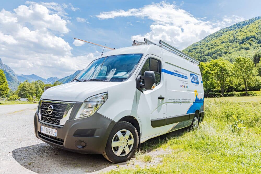 Camping-car blanc et bleu avec antenne déployée, garé au bord d’une route en pleine nature, face à des montagnes verdoyantes sous un ciel nuageux.
