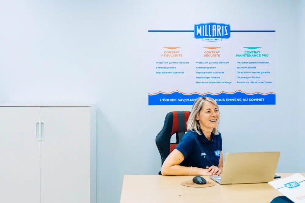 Employée en tenue bleue assise à un bureau, souriante devant un écran, avec une bannière murale “Millaris” et des offres de contrats (contrat régularité, sécurité et main’enance pro) en arrière-plan.