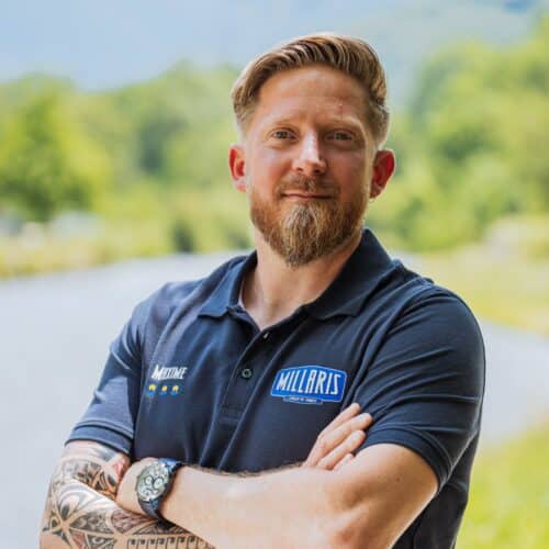 Portrait d’un homme, bras croisés, portant un polo bleu avec logos, devant un paysage vert flou et un ciel nuageux.