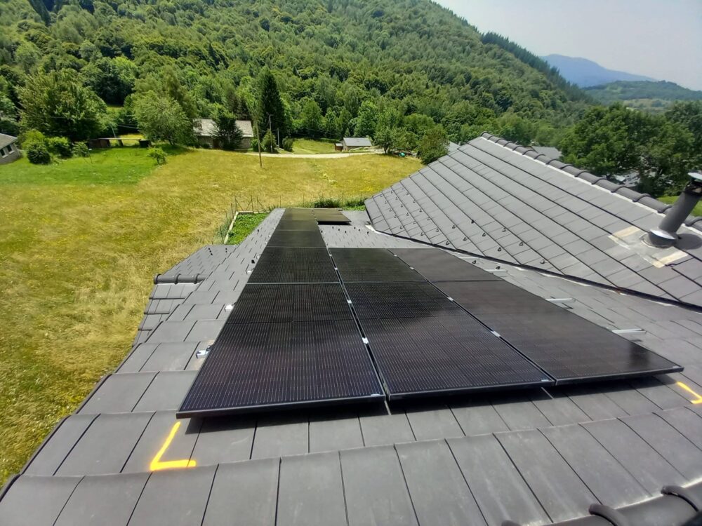 Toit couvert de panneaux solaires avec rangées de modules noirs et blancs, surplombant une prairie verte et des maisons au pied d’une colline boisée.