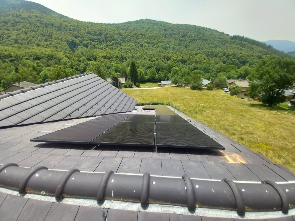 Toit équipé de panneaux solaires sur une maison, vue sur une pelouse et un village au pied de collines verdoyantes et de montagnes.