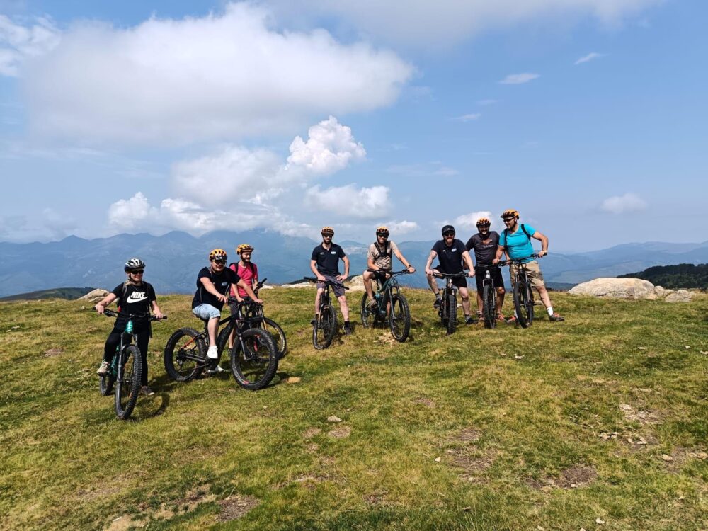 Groupe de sept cyclistes VTT en tenue de protection, posant au sommet d’une colline sur une herbe verte, avec des vélos tout-terrain et un paysage de montagnes en arrière-plan sous un ciel nuageux.