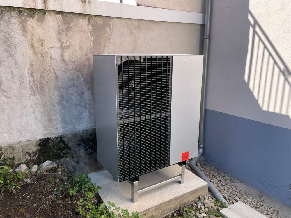 Unité extérieure de chauffage Vitmann avec grilles noires et carénage blanc posée sur une base en béton, contre un mur beige avec tuyauteries métalliques et ombres projetées.