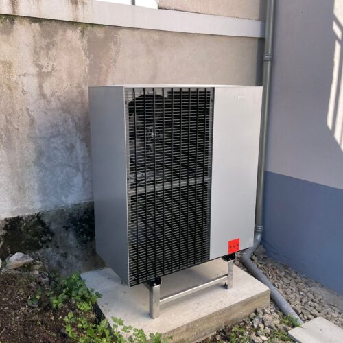 Unité extérieure de chauffage Vitmann avec grilles noires et carénage blanc posée sur une base en béton, contre un mur beige avec tuyauteries métalliques et ombres projetées.