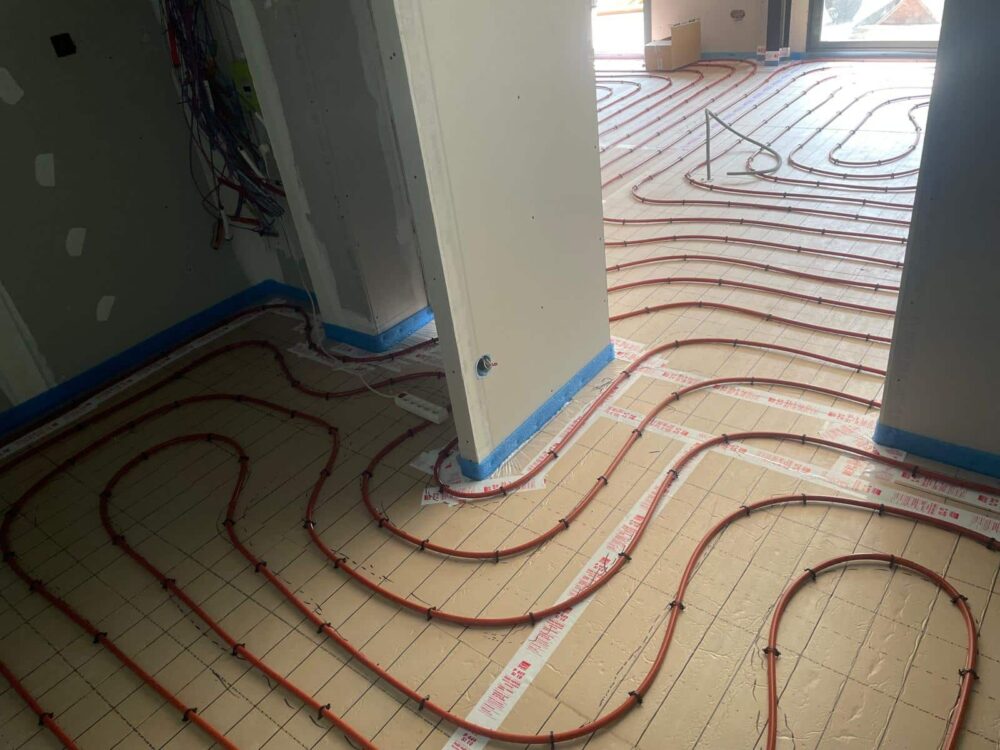 Plancher chauffant avec tuyaux rouges disposés en boucles sur un sol carrelé, vue depuis le passage entre des murs blancs dans une pièce en construction.
