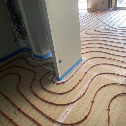 Plancher chauffant avec tuyaux rouges disposés en boucles sur un sol carrelé, vue depuis le passage entre des murs blancs dans une pièce en construction.