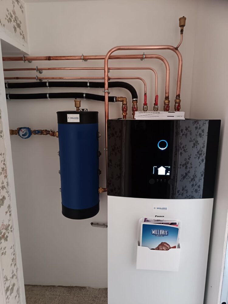 Chaudière avec tuyauterie en cuivre et ballon d’eau chaude bleu, dans une chaufferie intérieure blanche, avec affiche du fabricant MILARIS et écran de contrôle LED allumé.
