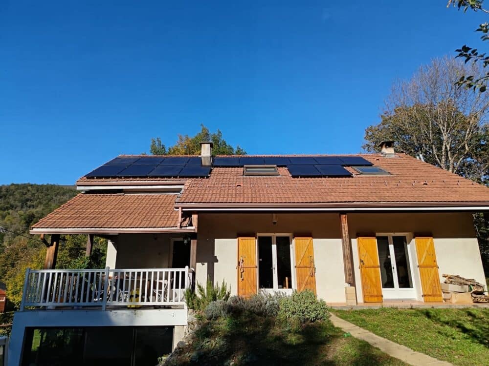 Maison individuelle avec toit en tuiles, panneaux solaires sur le versant et deux grandes portes-fenêtres aux volets jaunes, sous un ciel bleu.