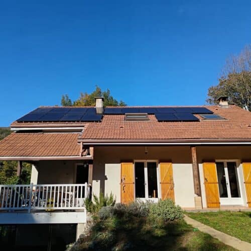 Maison individuelle avec toit en tuiles, panneaux solaires sur le versant et deux grandes portes-fenêtres aux volets jaunes, sous un ciel bleu.
