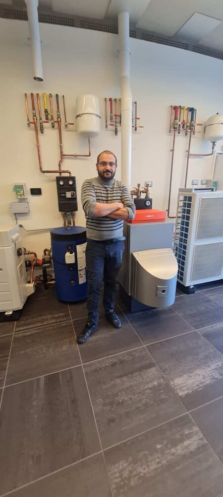 Technicien en salle de chauffage, debout avec les bras croisés devant une installation de chaudière et de tuyauterie industrielle, à côté d’une pompe à chaleur et d’équipements électriques.