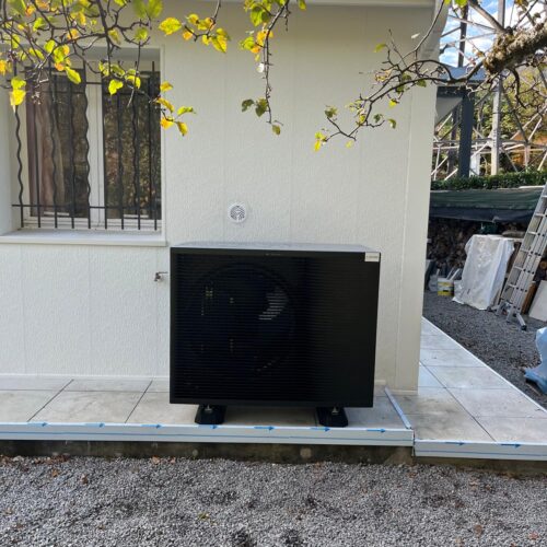 Climatiseur extérieur noir installé sur une plateforme au pied d’un mur blanc, avec des câbles bleus d’isolation, branché dans un jardin, sous des branches de feuilles vertes.