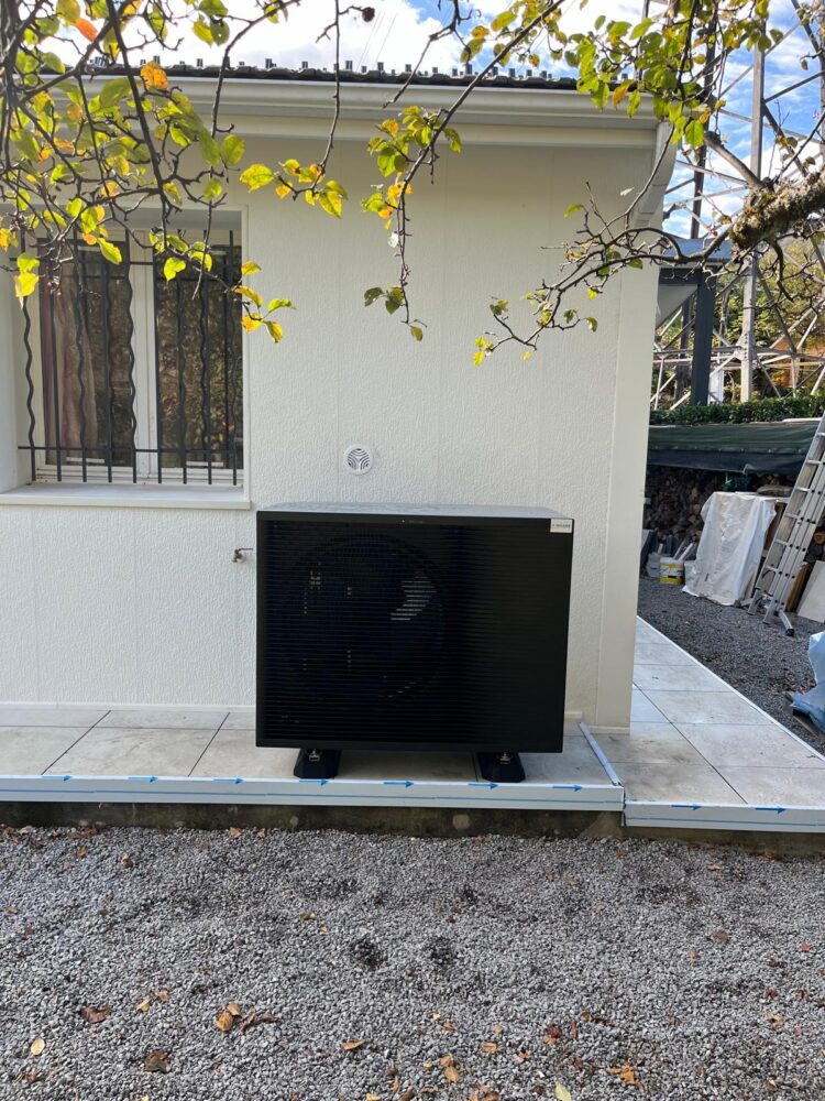 Climatiseur extérieur noir sur une dalle, adossé à un mur blanc, avec tuyaux bleus et branches de lierre vertes au premier plan.