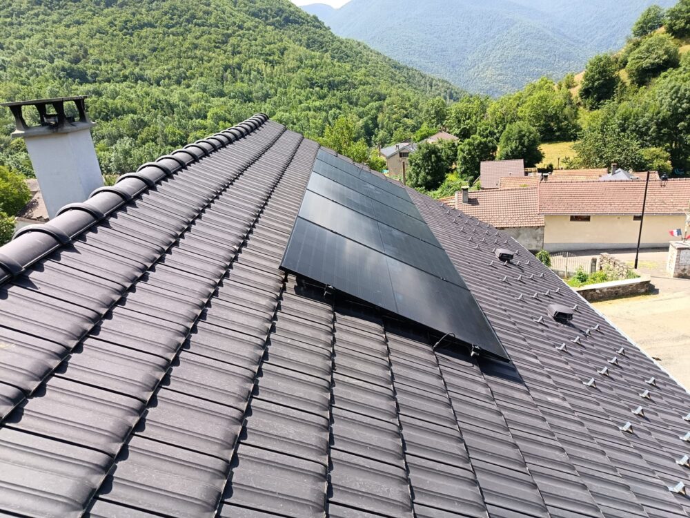 Toit en tuiles avec rangées de panneaux solaires photovoltaïques, vue depuis le haut d’une maison avec village et montagnes boisées en arrière-plan.