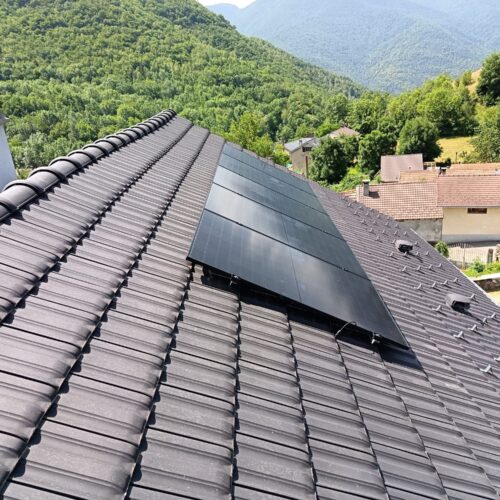 Toit en tuiles avec rangées de panneaux solaires photovoltaïques, vue depuis le haut d’une maison avec village et montagnes boisées en arrière-plan.