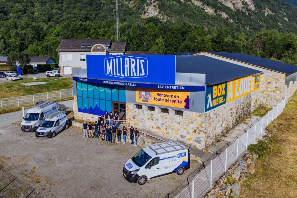 Façade d’un bâtiment « MILHARI » avec fourgons de l’entreprise et une équipe debout devant, en arrière-plan une colline couverte de végétation et un paysage montagneux.