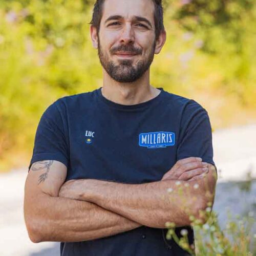 Portrait d’un homme souriant, bras croisés, portant un t-shirt bleu avec un badge « MILLARIS », en extérieur devant une végétation verte floue.