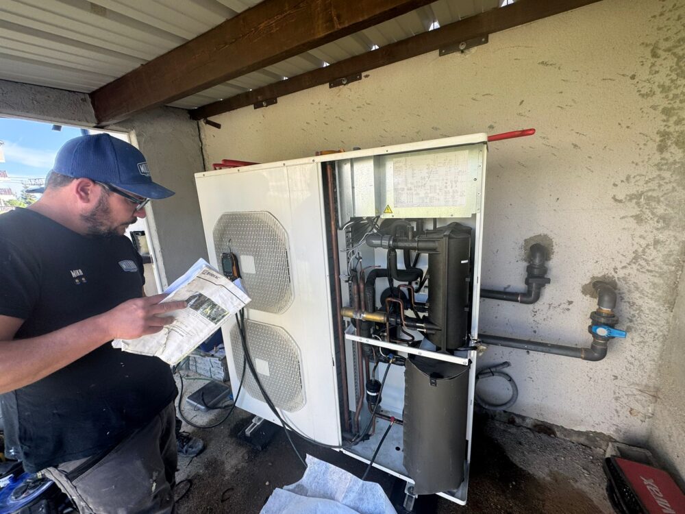 Technicien en uniforme bleu examinant une documentation près d’une pompe à chaleur extérieure, avec l’armoire ouverte montrant des tuyaux et composants électriques.
