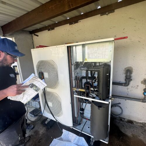 Technicien en uniforme bleu examinant une documentation près d’une pompe à chaleur extérieure, avec l’armoire ouverte montrant des tuyaux et composants électriques.