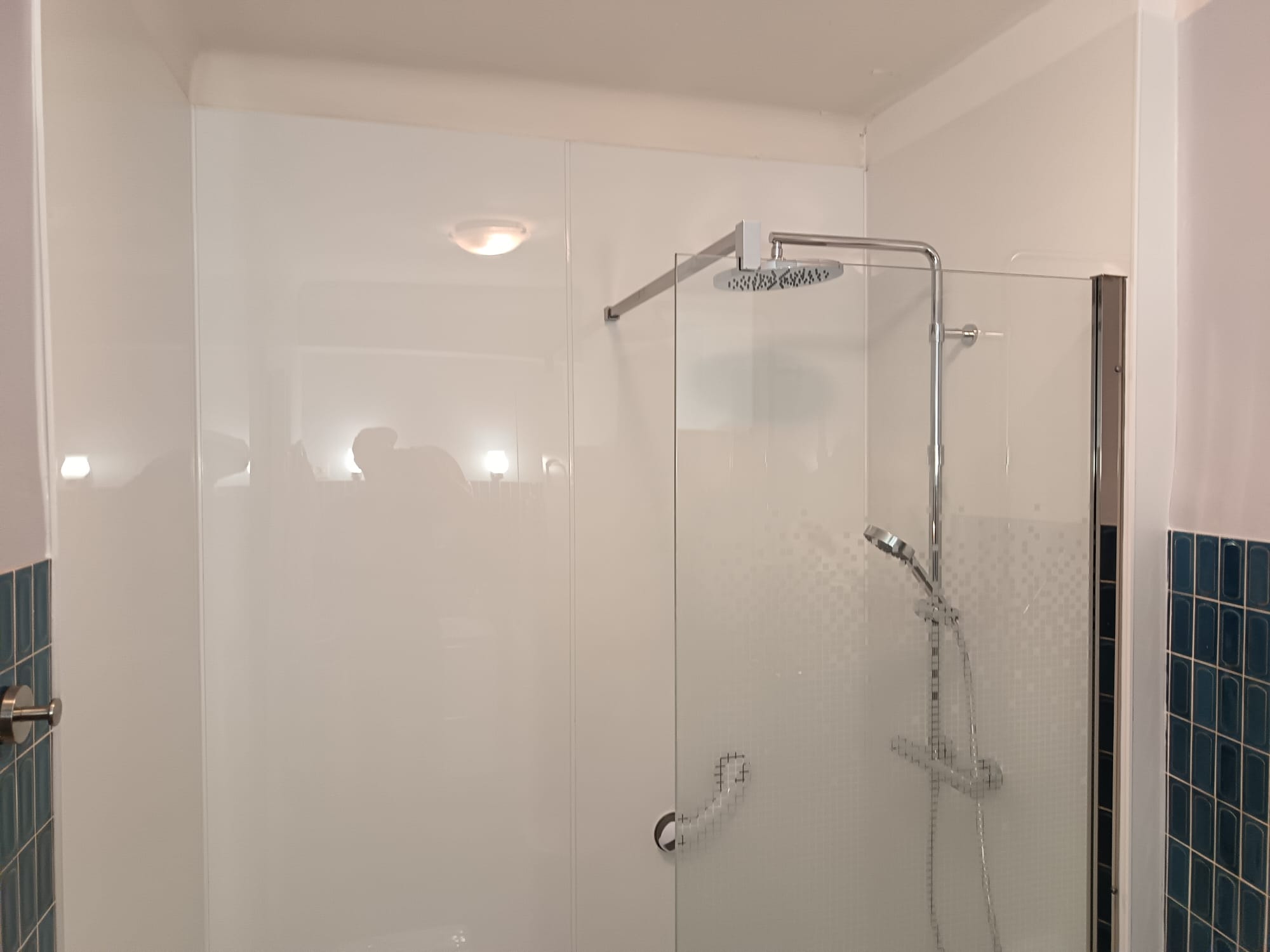 Douche à l’italienne avec paroi vitrée, pommeau de douche chromé et flexible, carreaux bleus au mur et éclairage au plafond.