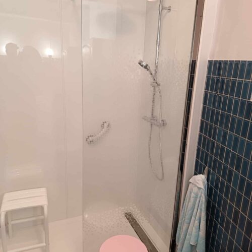 Douche vitrée avec pommeau de douche au plafond, mur carrelé bleu et rideau de douche, vue depuis la salle de bain avec toilettes et serviette visibles