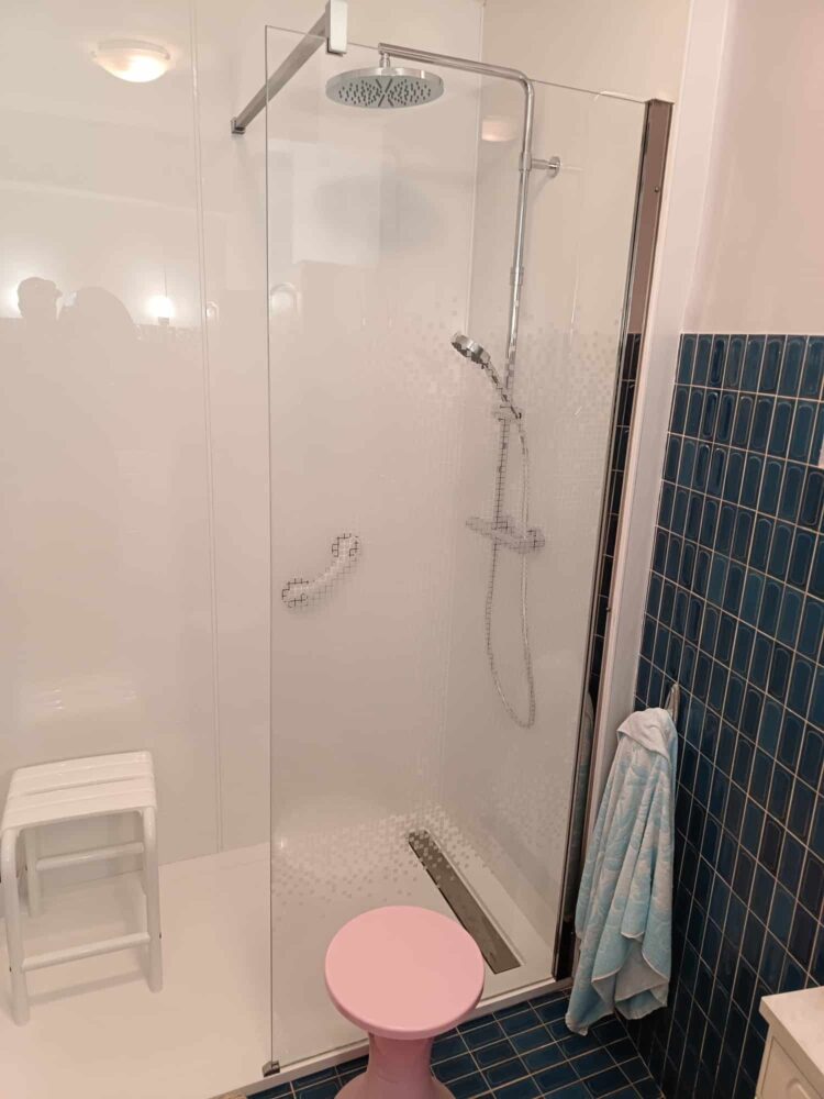 Douche vitrée avec pommeau de douche au plafond, mur carrelé bleu et rideau de douche, vue depuis la salle de bain avec toilettes et serviette visibles