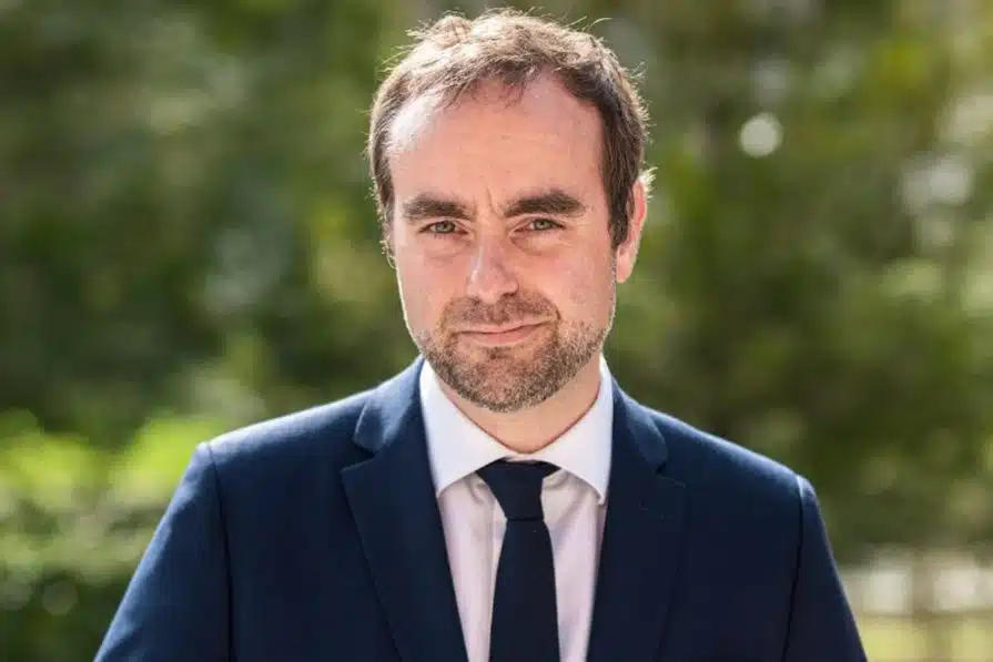 Portrait d’un homme d’affaires portant un costume bleu et une cravate noire, sur fond de jardin vert flou.