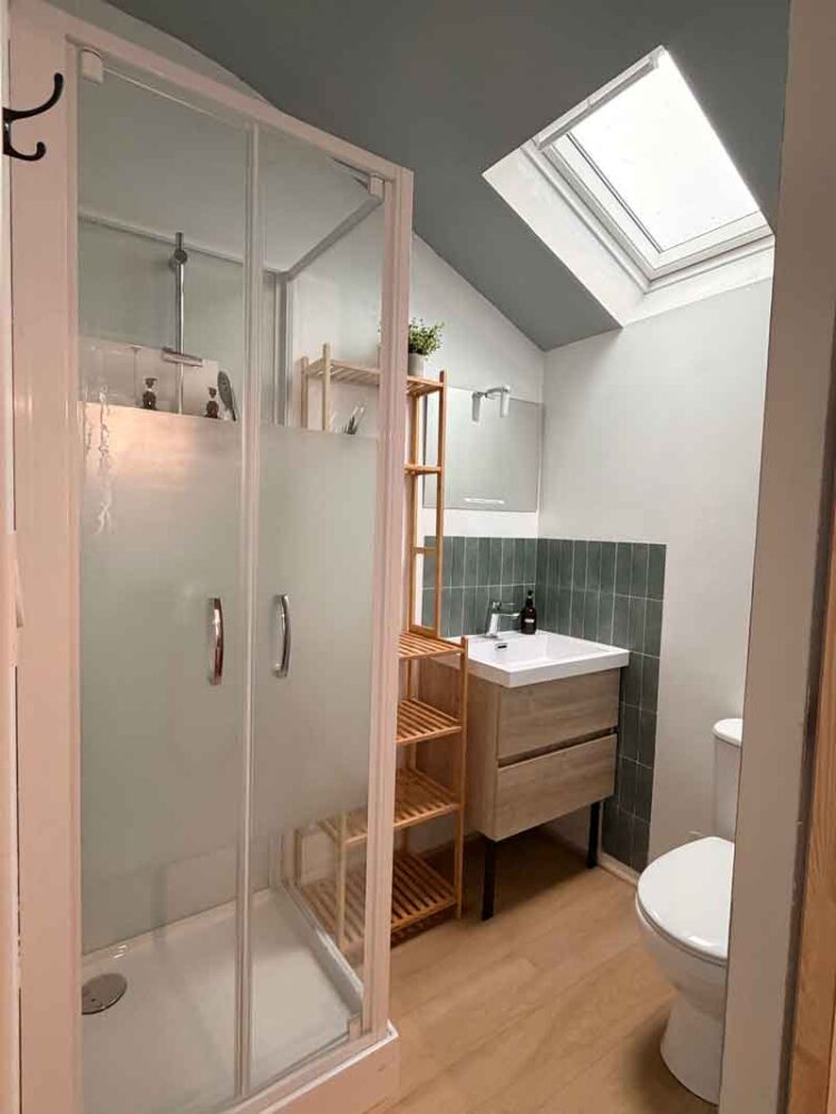 Salle de bain moderne avec douche vitrée, meuble vasque en bois, petite fenêtre de toit éclairant la pièce et WC au premier plan.