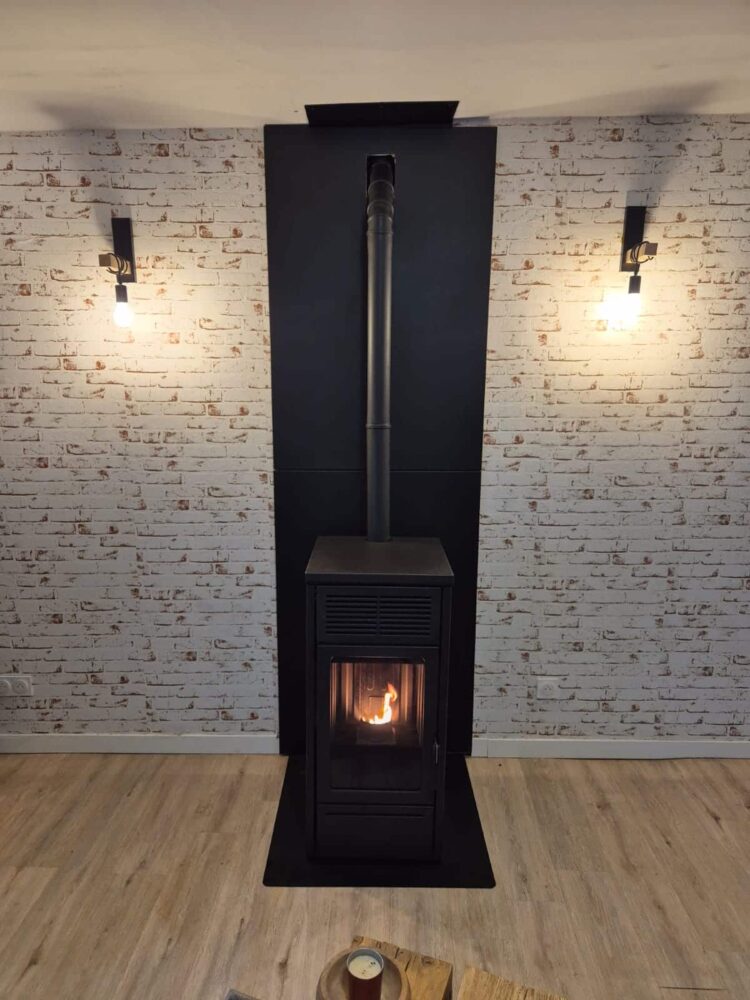 Poêle à bois noir avec conduit central et flamme visible dans la chambre, adossé à un mur en briques blanches, avec deux appliques murales allumées et un sol en parquet.