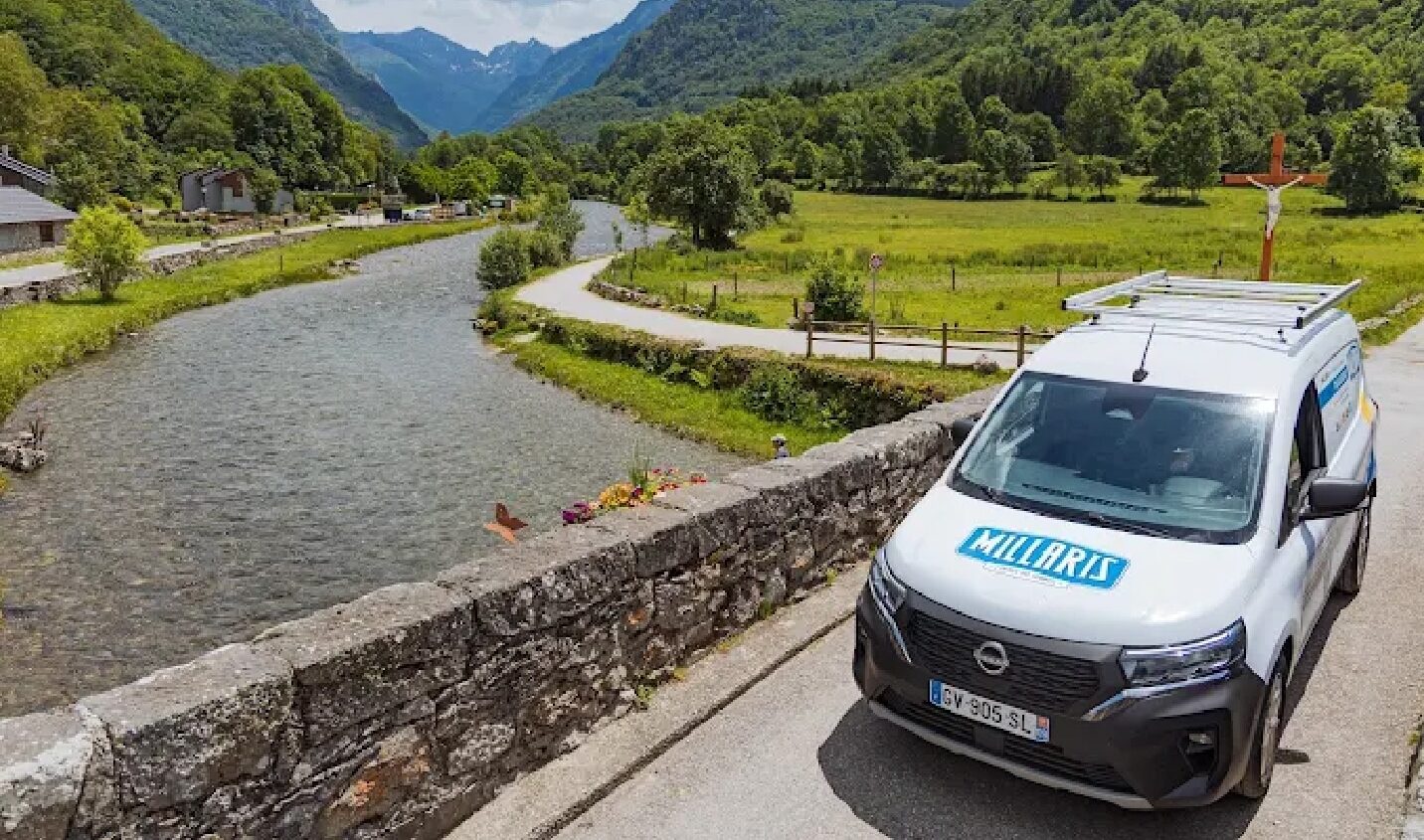 Véhicule blanc type ambulance garé sur un chemin près d’une rivière, dans une vallée verdoyante entourée de montagnes.
