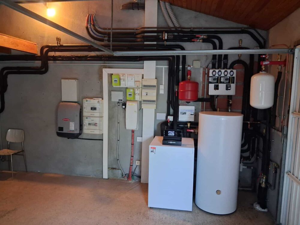 Local technique de chauffage avec tuyauteries en cuivre noires, armoire électrique, ballon d’eau chaude blanc, vase rouge et groupes de régulation montés au mur.