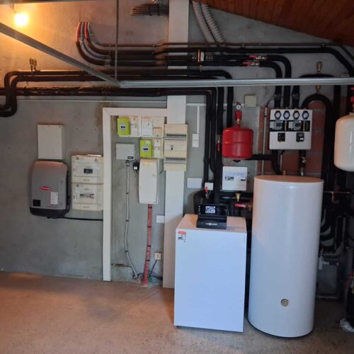 Local technique de chauffage avec tuyauteries en cuivre noires, armoire électrique, ballon d’eau chaude blanc, vase rouge et groupes de régulation montés au mur.